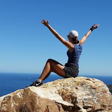 woman sits at summit on rock, arms in air, ocean and sky beyond
