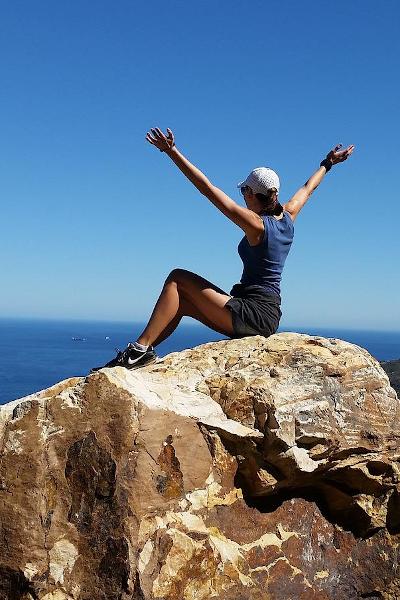 woman sits at summit on rock, arms in air, ocean and sky beyond